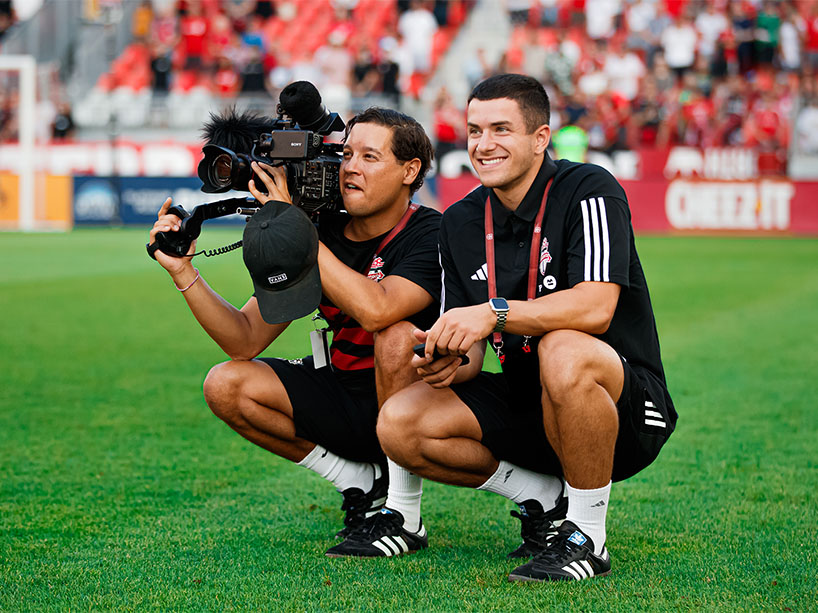 Two young men, one with a video camera in hand, kneel on the side of the field at a TFC soccer game. 